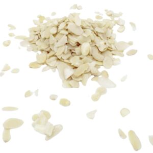 Scattered on a white surface is a stack of Sliced Peeled Almonds. The thin slices of light cream-colored almonds vary in size and have a slightly shiny texture. Some slices are piled together while others are scattered around the central stack.