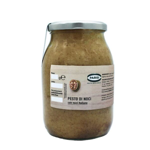 L'immagine mostra un vasetto di vetro di Pesto di Noci, un pesto di noci. Il barattolo ha un coperchio in metallo e un'etichetta beige con testo verde e nero. L'etichetta presenta anche un piccolo logo circolare con l'illustrazione di una noce e include ingredienti in italiano. Il contenuto appare cremoso con una consistenza grossa.